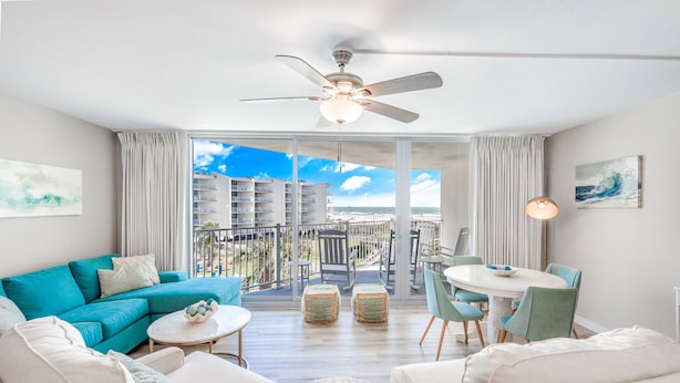 A modern living room with a balcony, a ceiling fan, and a dining area with chairs and a table. - Sandcastle Condominium Unit 329 (Port Aransas)