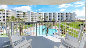 A balcony with white chairs overlooking a swimming pool and a building with multiple balconies. - Sandcastle Condominium Unit 329 (Port Aransas)