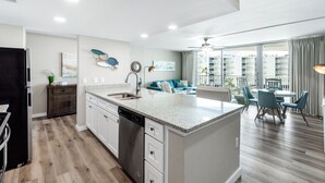 A modern kitchen with a dishwasher, a dining area with a round table and chairs, and a balcony with a view of the city. - Sandcastle Condominium Unit 329 (Port Aransas)