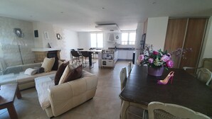 Amplia sala de estar con sofá, mesa de comedor y sillas. Se observa una cocina al fondo, junto con un comedor.