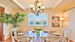A dining area with a wooden table, white chairs, a chandelier, a window with a view, and a potted plant. - On The Beach Clubhouse - Bayfront beauty (San Diego)