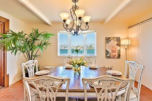 A dining area with a wooden table, white chairs, a chandelier, a window with a view, and a potted plant.