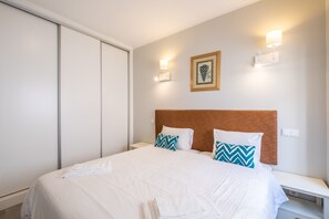 Habitación de hotel con cama, ropa blanca, dos almohadas con diseño de chevron, mesita de noche, un cuadro enmarcado en la pared y una lámpara fijada a la pared.