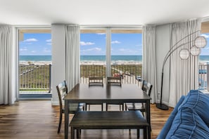 A spacious room with a large dining table, a blue sofa, and a view of the beach and ocean through large windows. - Sandcastle Condominium Unit 438 (Port Aransas)