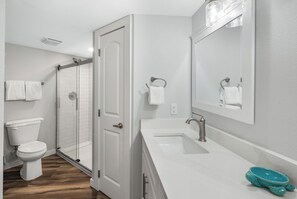 A bathroom with a white toilet, a glass-enclosed shower, a white sink with a mirror, and a towel rack. - Sandcastle Condominium Unit 438 (Port Aransas)
