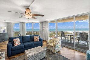 A spacious living room with a large blue sofa, a patterned armchair, and a ceiling fan. A balcony with a view of the ocean is visible through large windows. - Sandcastle Condominium Unit 438 (Port Aransas)