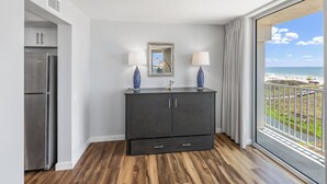 A modern room with a dark wooden cabinet, a stainless steel refrigerator, and a large window with a view of the beach. - Sandcastle Condominium Unit 438 (Port Aransas)