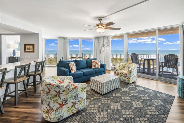 A spacious living room with a blue sofa, matching armchairs, a coffee table, and a view of the ocean. - Sandcastle Condominium Unit 438 (Port Aransas)