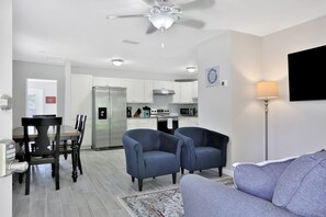 A modern living room with a gray sofa, two blue armchairs, a dining table with chairs, and a kitchen in the background.