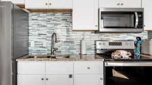 A modern kitchen with white cabinets, a stainless steel refrigerator, a microwave, and a sink with a mosaic backsplash. - Sandcastle Condominium Unit 506 (Port Aransas)