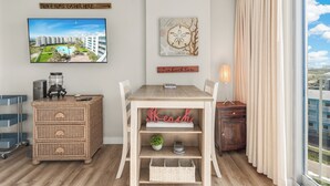A modern living room with a wooden dining table, wicker side table, and a view of a pool and buildings through large windows. - Sandcastle Condominium Unit 506 (Port Aransas)