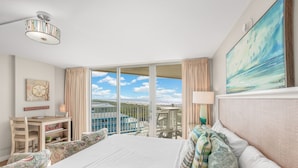 A hotel room with a large bed, a desk, a chair, and a view of the sea through large windows. - Sandcastle Condominium Unit 506 (Port Aransas)