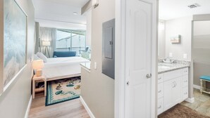 A hotel room with a bed, a small bedside table with a lamp, a bathroom with a sink and mirror, and a view of the outside balcony.