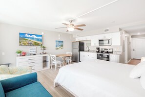 Un dormitorio moderno con una cama grande, un escritorio con silla, kitchenette y ventilador de techo.