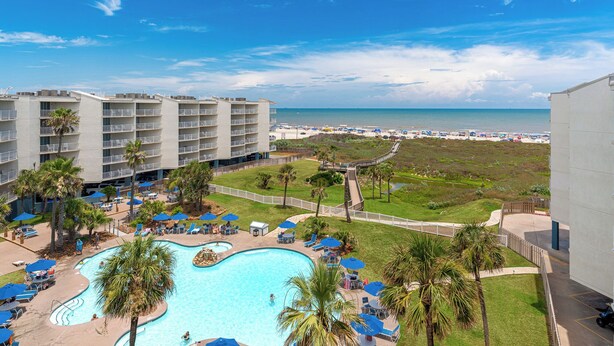 A hotel with a swimming pool surrounded by lounge chairs and umbrellas, a beach in the background, and a clear sky.