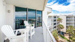 A balcony with white Adirondack chairs and a table, overlooking a beach and other buildings.