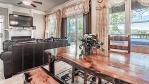 A dining area with a wooden table, a black leather sofa, a television, and a view of greenery outside.