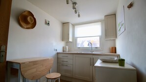 Een kleine keuken met witte kastjes, een houten kruk, een hoed aan de muur en een raam.