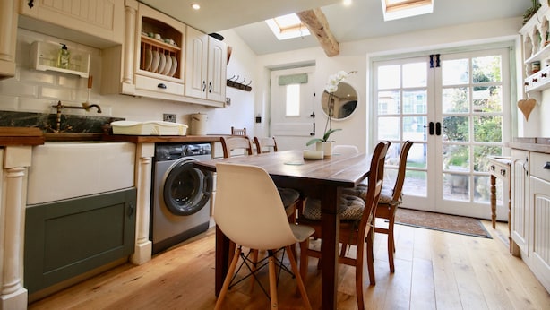 Een keuken met houten kastjes, een wasmachine, een eettafel met stoelen en een groot raam.