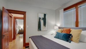 Un dormitorio con una cama grande, una mesita de noche con lámpara, un cuadro en la pared y una ventana con persianas.