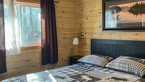 Een houten kamer met een bed, een raam met gordijnen, een wandlamp en een lijst met een boslandschap.