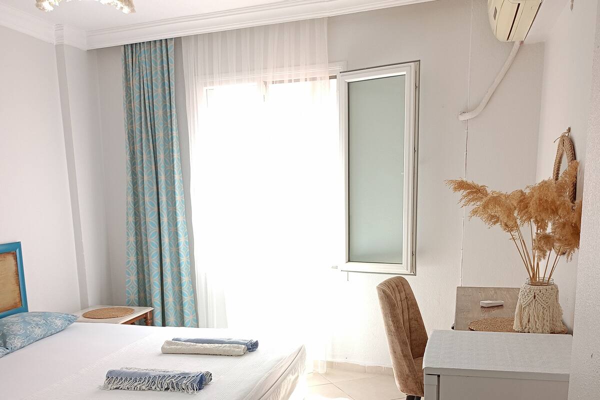 A bedroom with a bed, a desk, a chair, a refrigerator, and a window with curtains.
