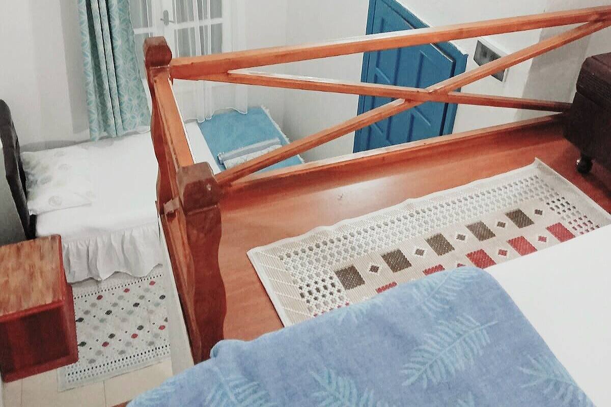 A small, simple room with a wooden bed, a small table, a chair, a window with curtains, and a wall-mounted air conditioner.