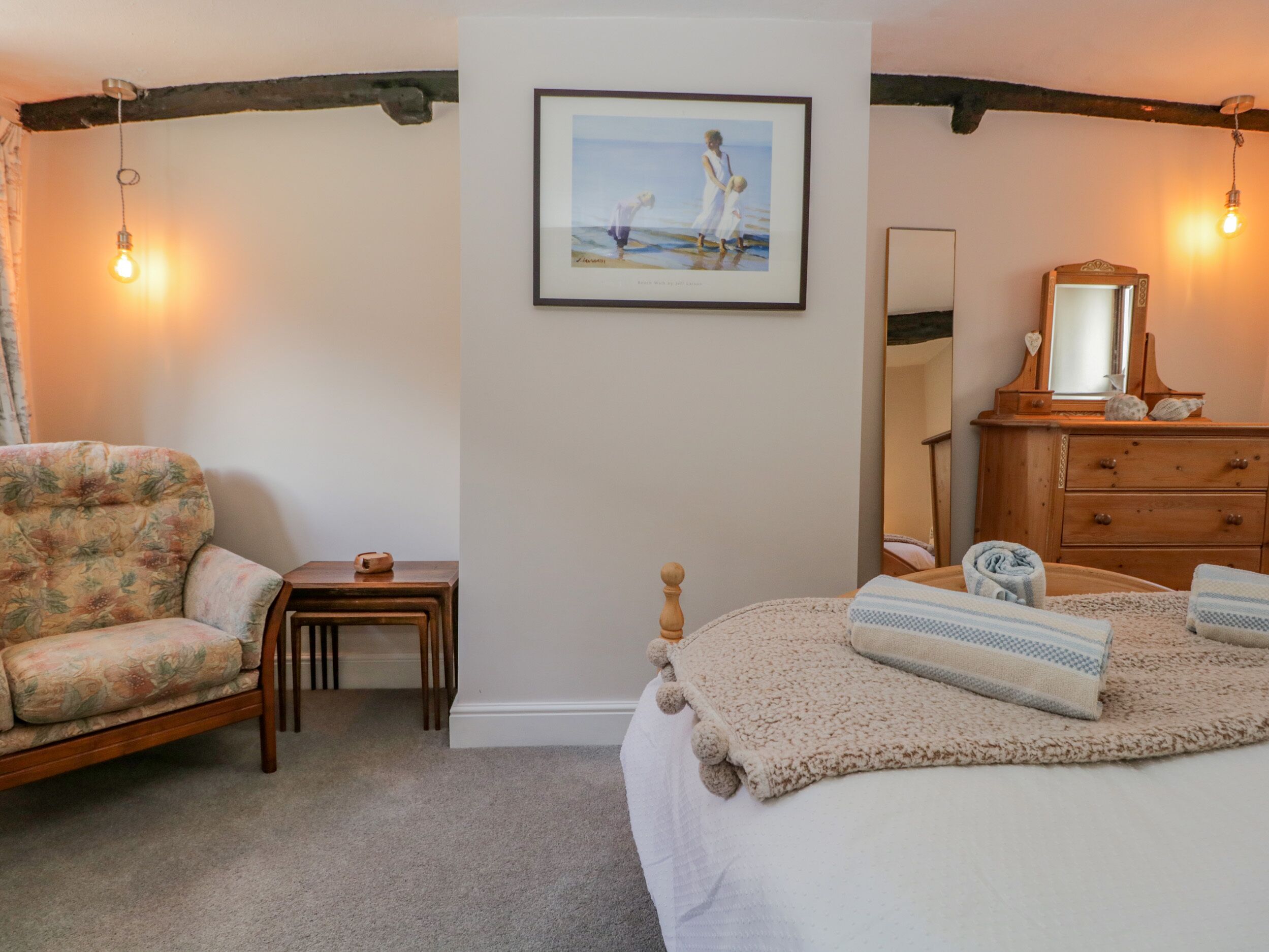 A bedroom with a bed, a chair, a small table, a dresser, a mirror, and a framed picture on the wall.