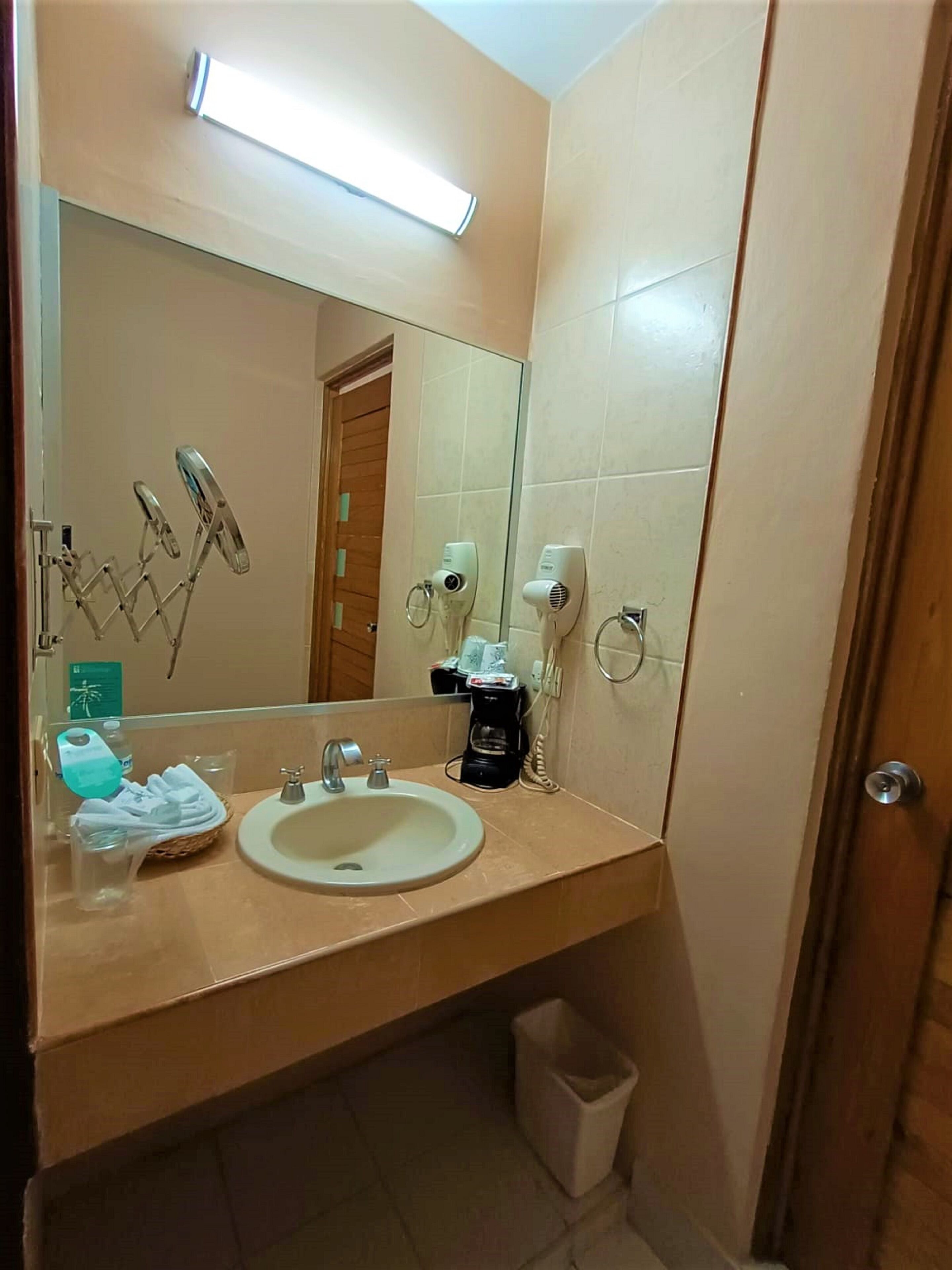 A bathroom with a sink, mirror, hairdryer, and a trash bin.