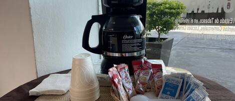 Une machine à café, une tasse et un panier contenant des sachets de café sont posés sur une table près d’une fenêtre.