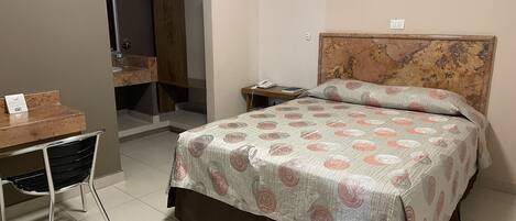 A hotel room with a bed, a desk, a chair, a nightstand, and a bathroom door.