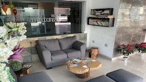 A modern living room with a grey sofa, ottomans, and a coffee table with magazines. There are potted plants and a wooden tree stump. - Hotel Santo Domingo (Villahermosa)