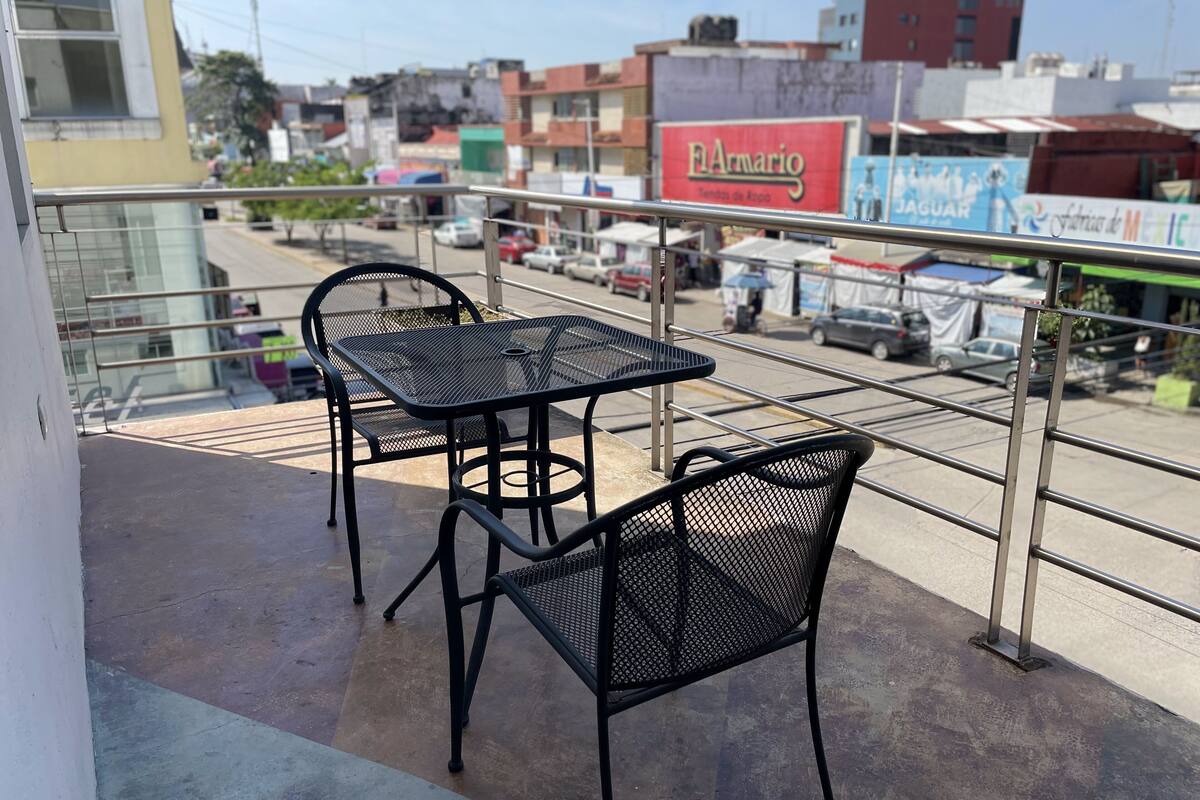 Un balcón con dos mesas y sillas de metal, con vista a una calle con varios edificios y coches estacionados.