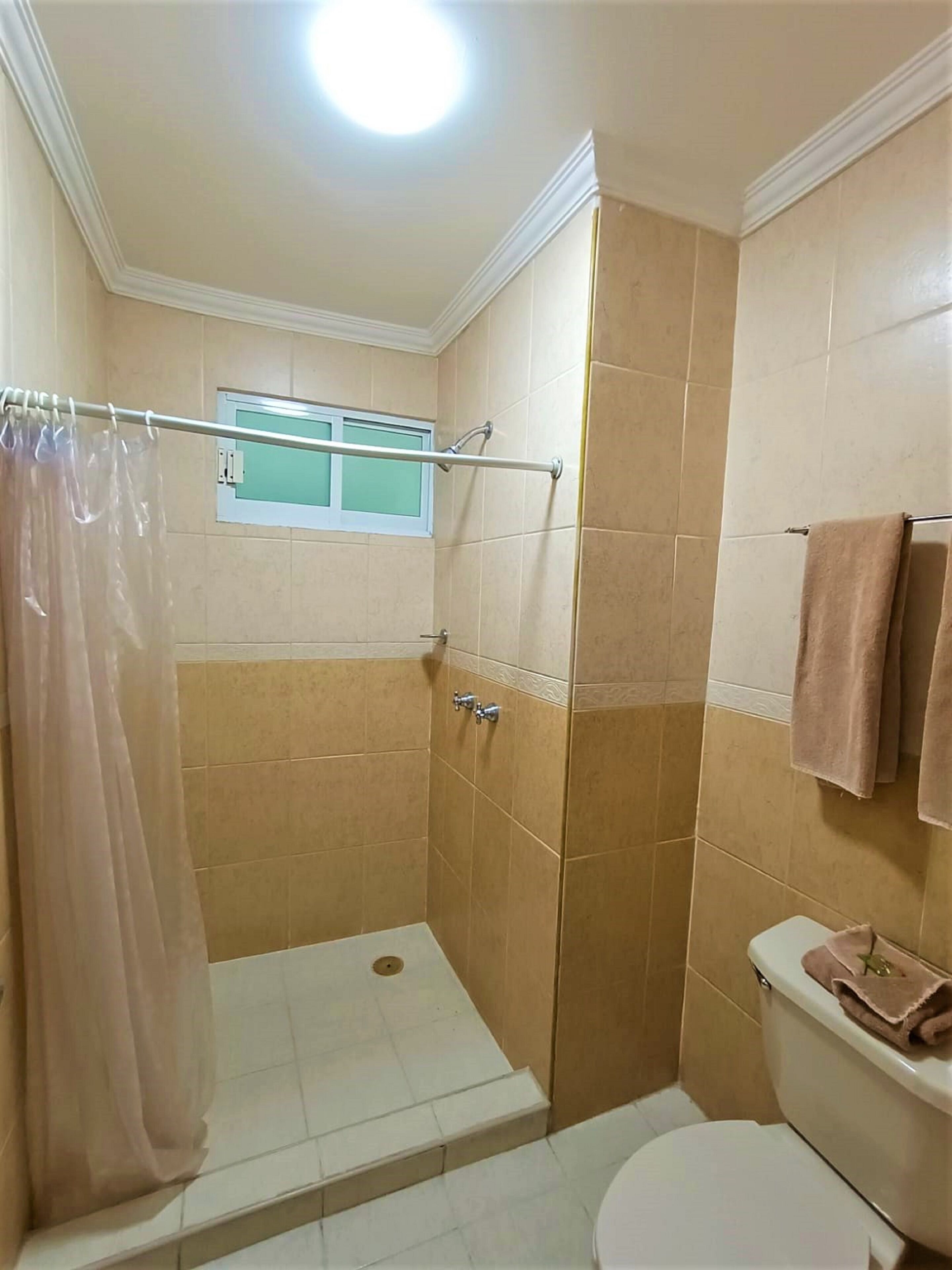 Une petite salle de bain carrelée, équipée d’une douche, de toilettes et d’une fenêtre.
