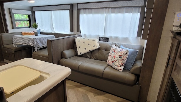 A compact RV interior with a kitchenette, a seating area with a sofa and cushions, and a window with curtains. - RV Glamping in the Heart of Downtown Eatonville with a Mountain View (Eatonville)