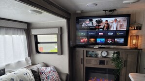 A cozy RV interior with a flat-screen TV mounted above a fireplace, a comfortable seating area, and a window with curtains. - RV Glamping in the Heart of Downtown Eatonville with a Mountain View (Eatonville)