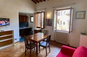 Un coin repas avec une table et des chaises en bois, une cuisine en arrière-plan, un canapé rouge et une fenêtre donnant sur des immeubles.