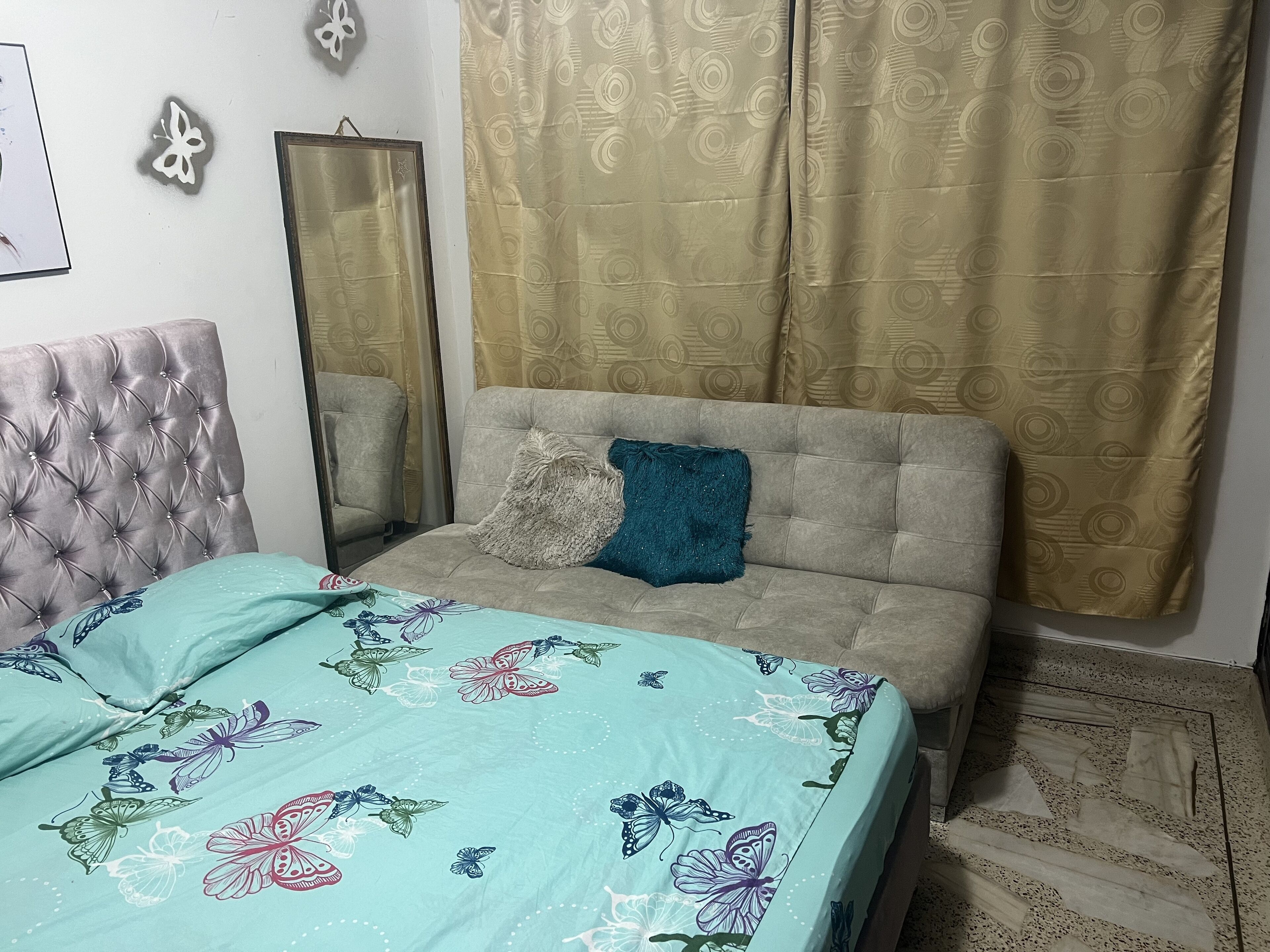 Ein Schlafzimmer mit einer gepolsterten Kopfteile, einer floralen Bettdecke, einem grauen Sofa, einem blauen Kissen und einem gemusterten Vorhang.