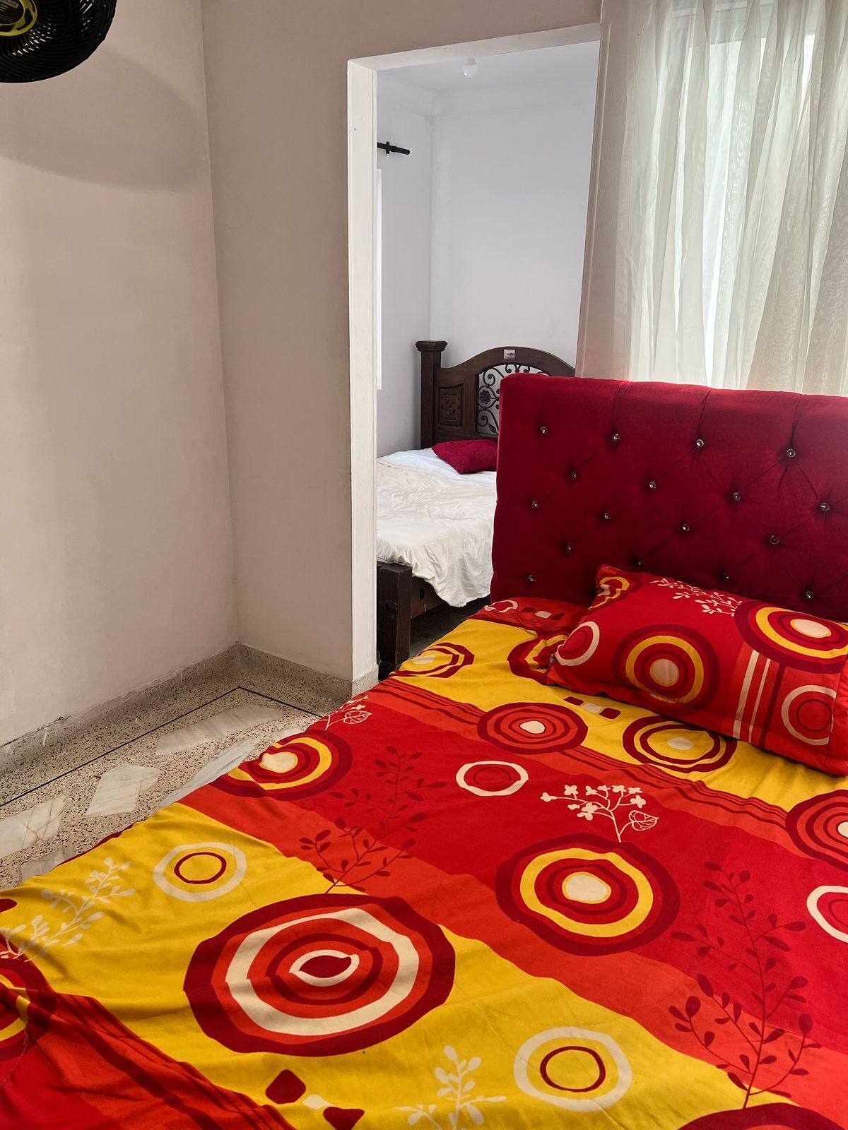 A bedroom with a bed covered in a colorful bedspread, a red upholstered headboard, and a wooden headboard bed in the background.