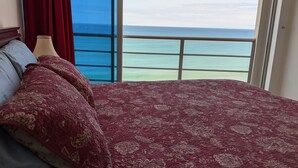 Un lit recouvert d’une couvre-lit à motifs rouges, avec un oreiller bleu et un oreiller rouge. Une fenêtre offre une vue sur l’océan.