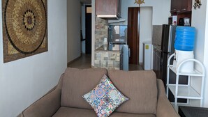 Un salon comprenant un canapé, un coussin aux couleurs vives, un coin cuisine avec un distributeur d’eau et un élément décoratif mural.