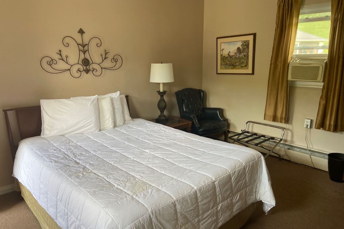 Une chambre d’hôtel avec un grand lit, une chaise, une table de chevet, une lampe, un tableau au mur et une fenêtre avec des rideaux.