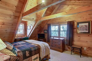 A cozy attic bedroom with a large bed, a dresser, a TV, and a window with a view of trees.