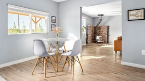 A modern dining area with a round table, white chairs, and a vase of flowers. A brick fireplace and a telescope are visible in the background.