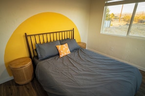 Una cama con cabecera, almohadas, una mesita de noche de madera, una cesta de mimbre y una ventana con vista al paisaje.