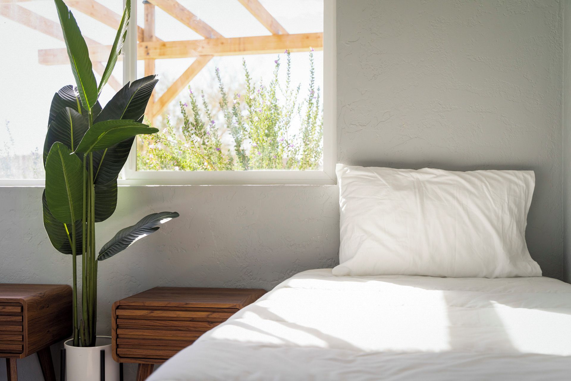 Un dormitorio con una cama, una ventana, una planta y mesitas de noche de madera.