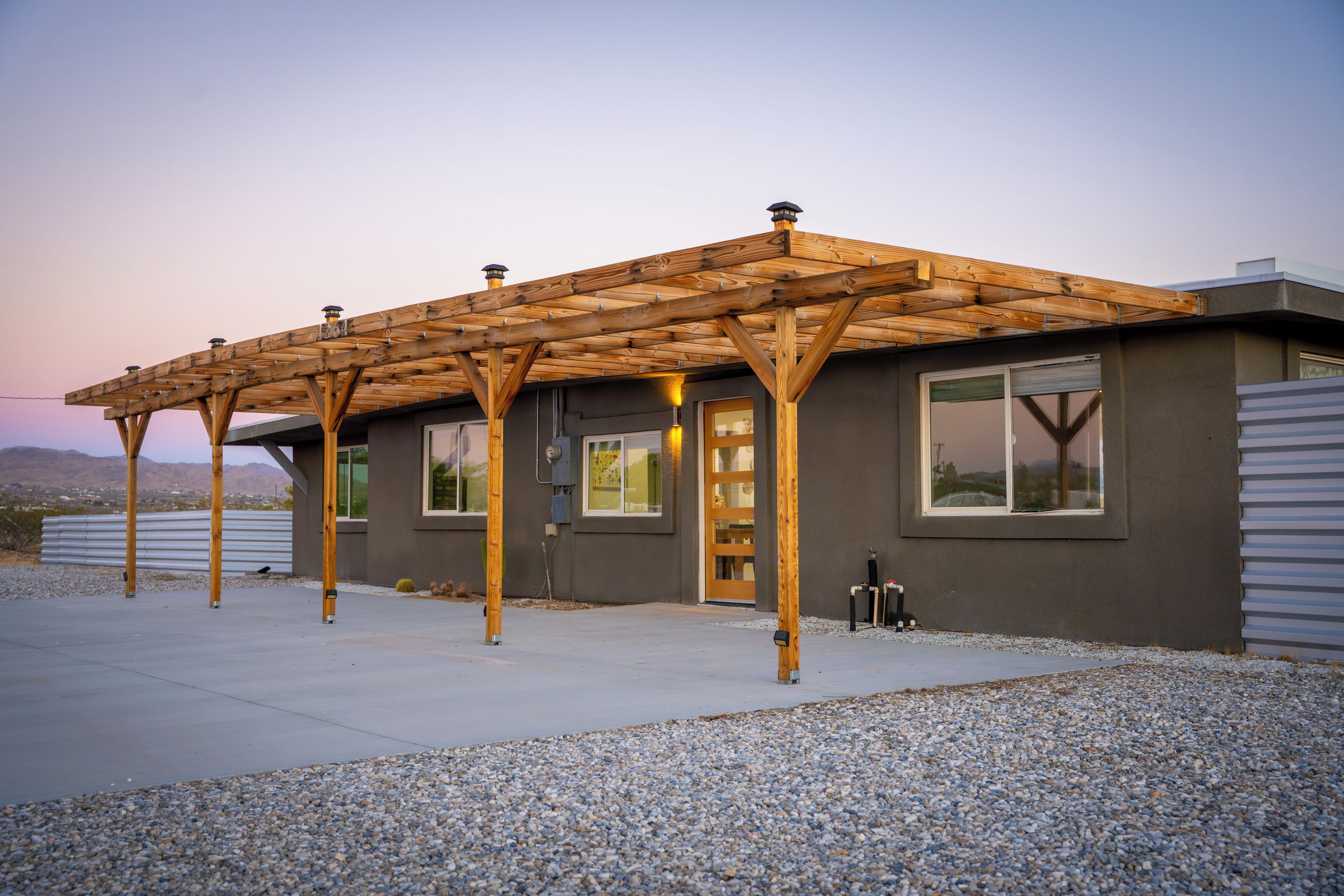 A modern house with a wooden pergola structure, large windows, and a gravel driveway.