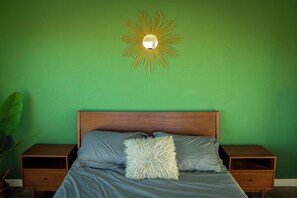 Ein Schlafzimmer mit grüner Wand, einem Holzkopfende, einem Bett mit grauer Bettwäsche und einem Nachttisch auf jeder Seite.