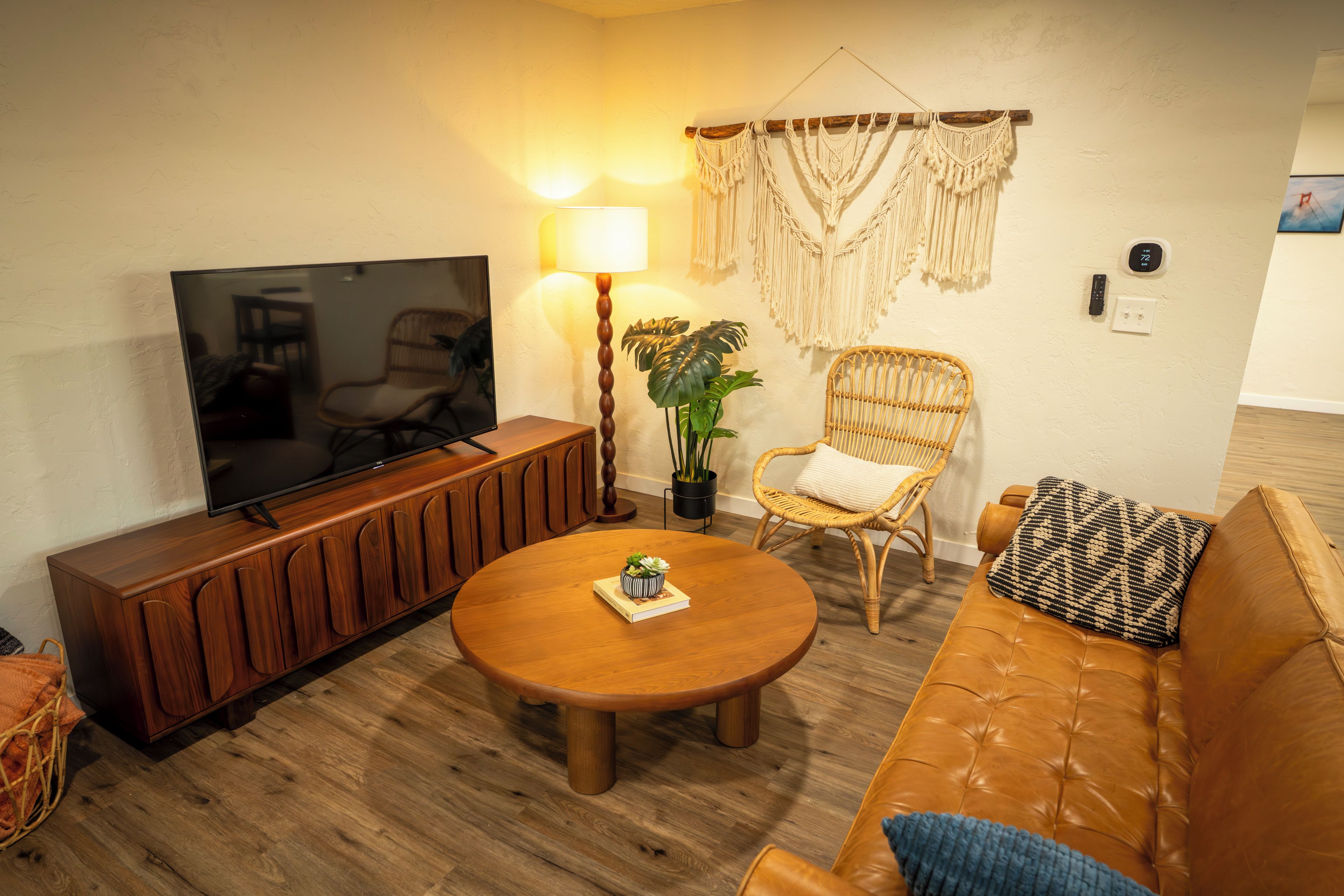A living room with a TV, wooden furniture, a round coffee table, a wicker chair, and a macrame wall hanging.
