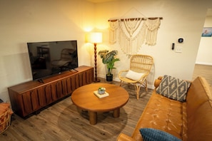 Una sala de estar con televisor, muebles de madera, una mesa de centro redonda, una silla de mimbre y un colgador de macramé en la pared.